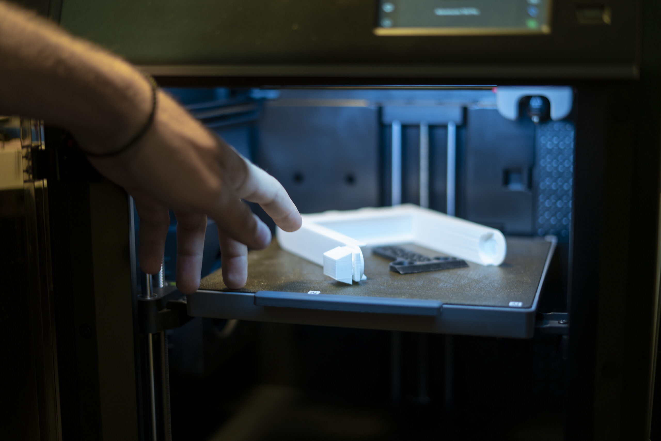A 3D printer is in action, producing a white item with intricate designs on a build plate in a well-lit workshop, showcasing advanced printing technology.